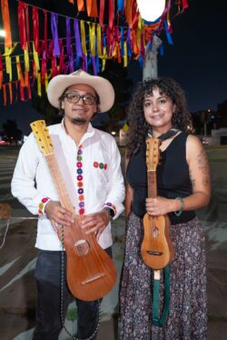 César and Rebecca are standing with their jaranas at the fandango at East Side Cafe. There are stringed ribbons as decorations along with lights in the background. Rebecca is wearing a black blouse and long skirt with earth tone patterns. César is wearing a white shirt with embroidery in the middle, hat, and black pants. Photo credit: IG @chicawithbees