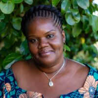 Muisi-kongo Malonga is pictured wearing a brightly colored blue and orange floral-patterned top. She is seated outdoors with a lush green background of leaves.