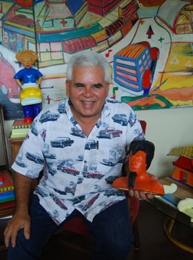Magu holds Indio During His Egyptian Period with Having a Car Baby (background left, created while volunteering at the women’s prison in Chino, California) and sitting in front of one of his Visiting Magulandia paintings.